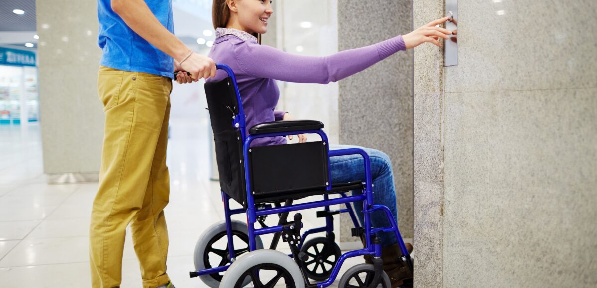 A person in a wheelchair reaches for an elevator button with assistance nearby. Modular Wheelchair Ramps for Sale.