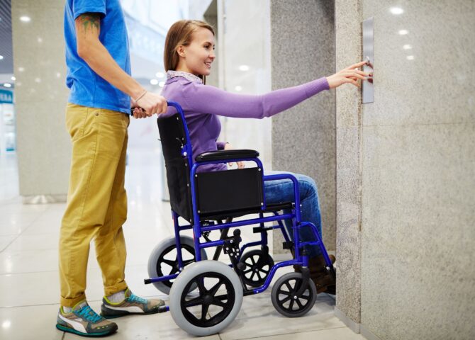 A person in a wheelchair reaches for an elevator button with assistance nearby. Modular Wheelchair Ramps for Sale.