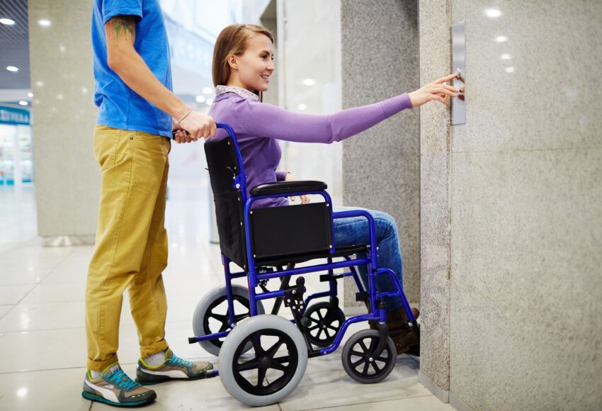 A person in a wheelchair reaches for an elevator button with assistance nearby. Modular Wheelchair Ramps for Sale.